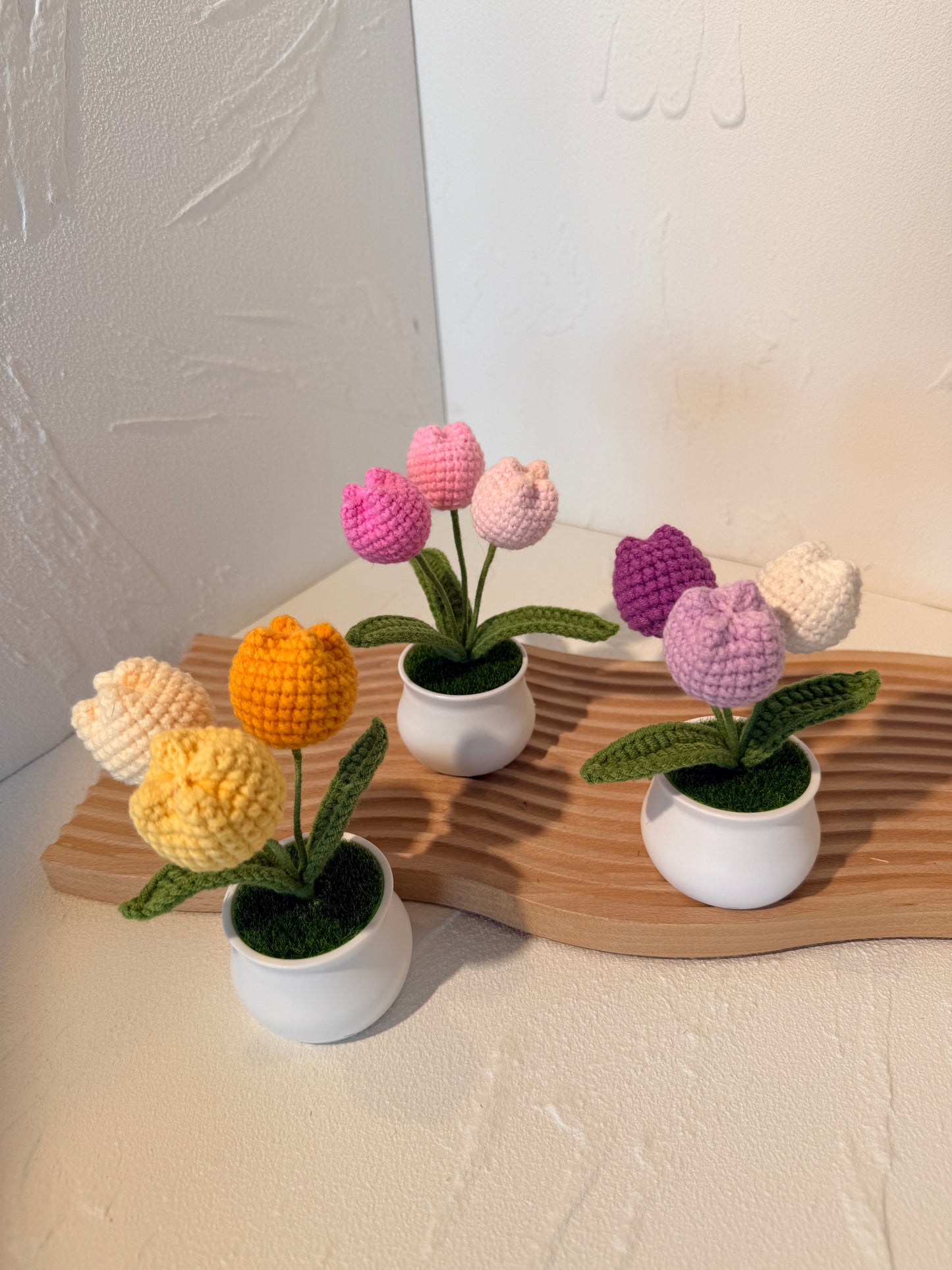 Colorful knitted flowers in white pots on a wooden board against a neutral wall.