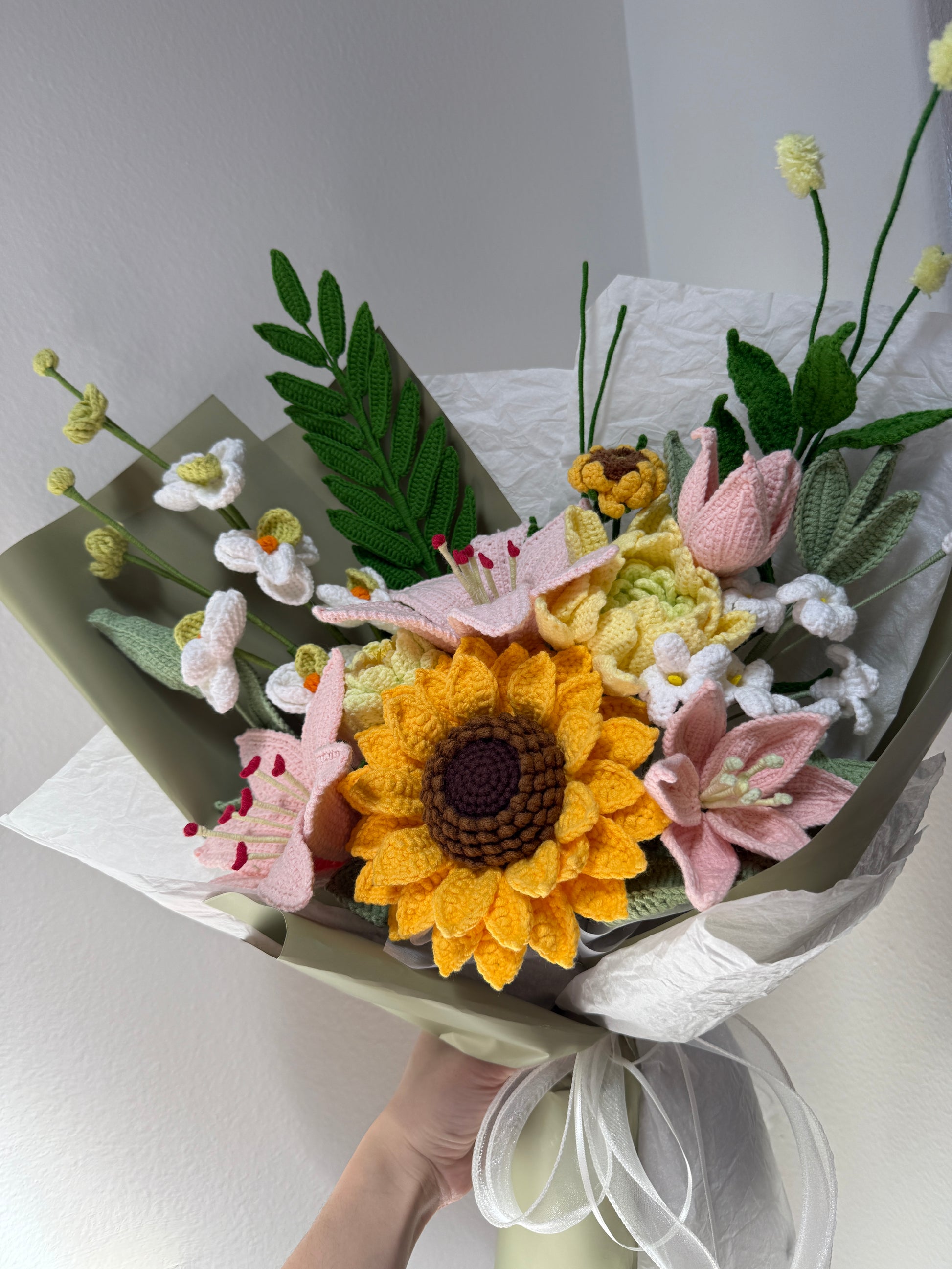 Bouquet of crocheted flowers including a sunflower, pink lilies, and green leaves on a white background.